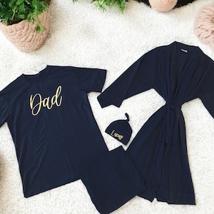 A flat-lay image featuring a navy blue maternity robe and pajama set with "Dad" written in gold script on a T-shirt, a matching robe, a baby hat with "Lucas" embroidered in gold, and a pair of pants, arranged on a soft white rug.