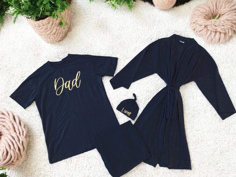A flat-lay image featuring a navy blue maternity robe and pajama set with "Dad" written in gold script on a T-shirt, a matching robe, a baby hat with "Lucas" embroidered in gold, and a pair of pants, arranged on a soft white rug.