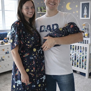 May include: A family poses in a nursery decorated with a space theme. The mother wears a navy blue robe with a space pattern. The father holds a baby wrapped in a matching navy blue space-themed swaddle. The father's shirt says "DAD".