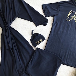 A flat-lay image featuring a navy blue maternity robe and pajama set with "Dad" written in gold script on a T-shirt, a matching robe, a baby hat with "Lucas" embroidered in gold, and a pair of pants, arranged on a soft white rug.