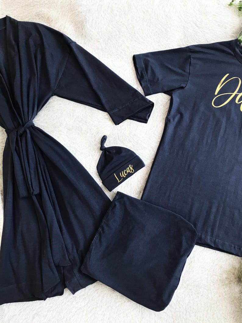 A flat-lay image featuring a navy blue maternity robe and pajama set with "Dad" written in gold script on a T-shirt, a matching robe, a baby hat with "Lucas" embroidered in gold, and a pair of pants, arranged on a soft white rug.