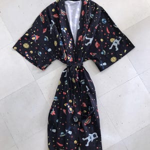 Family-themed clothing set featuring white T-shirts with space-themed "DAD," "BIG BRO," and "BIG SIS" text, a navy robe with colorful space designs, and a matching space-themed swaddle with a personalized hat. Laid out on a tiled floor.