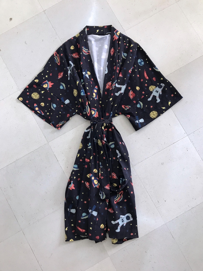 Family-themed clothing set featuring white T-shirts with space-themed "DAD," "BIG BRO," and "BIG SIS" text, a navy robe with colorful space designs, and a matching space-themed swaddle with a personalized hat. Laid out on a tiled floor.