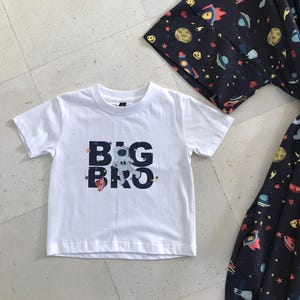 Family-themed clothing set featuring white T-shirts with space-themed "DAD," "BIG BRO," and "BIG SIS" text, a navy robe with colorful space designs, and a matching space-themed swaddle with a personalized hat. Laid out on a tiled floor.