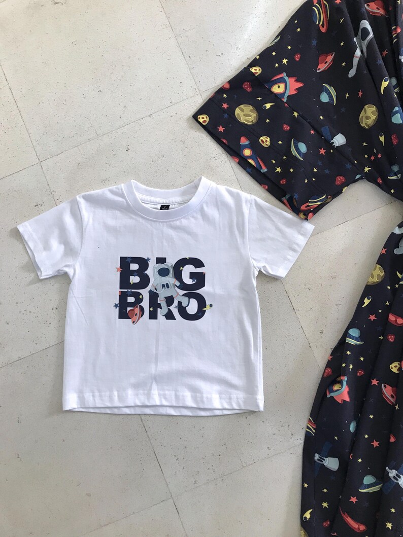 Family-themed clothing set featuring white T-shirts with space-themed "DAD," "BIG BRO," and "BIG SIS" text, a navy robe with colorful space designs, and a matching space-themed swaddle with a personalized hat. Laid out on a tiled floor.