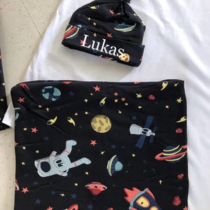Family-themed clothing set featuring white T-shirts with space-themed "DAD," "BIG BRO," and "BIG SIS" text, a navy robe with colorful space designs, and a matching space-themed swaddle with a personalized hat. Laid out on a tiled floor.
