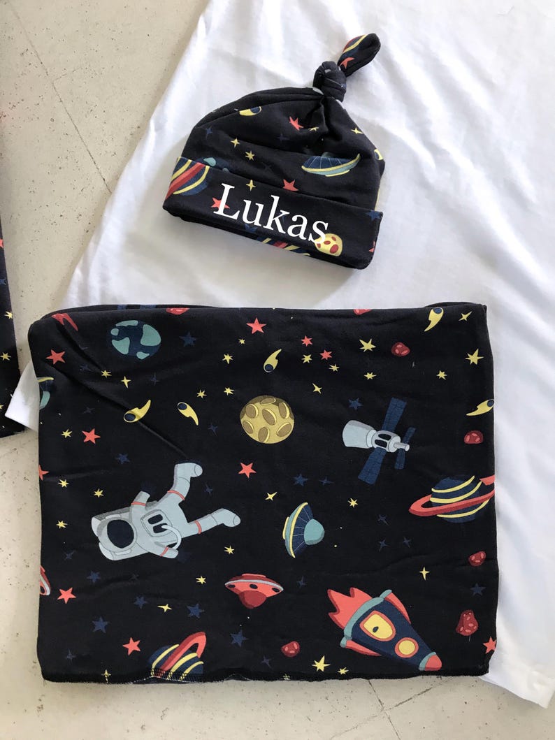 Family-themed clothing set featuring white T-shirts with space-themed "DAD," "BIG BRO," and "BIG SIS" text, a navy robe with colorful space designs, and a matching space-themed swaddle with a personalized hat. Laid out on a tiled floor.