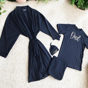A flat-lay image featuring a navy blue maternity robe and pajama set with "Dad" written in gold script on a T-shirt, a matching robe, a baby hat with "Lucas" embroidered in gold, and a pair of pants, arranged on a soft white rug.