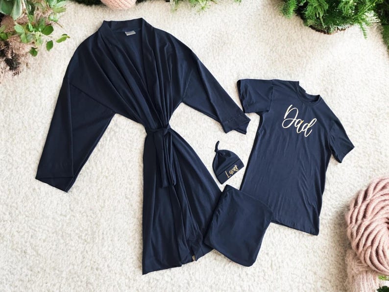 A flat-lay image featuring a navy blue maternity robe and pajama set with "Dad" written in gold script on a T-shirt, a matching robe, a baby hat with "Lucas" embroidered in gold, and a pair of pants, arranged on a soft white rug.
