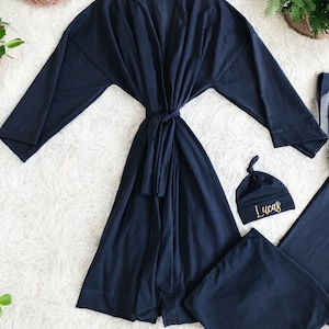 A flat-lay image featuring a navy blue maternity robe and pajama set with "Dad" written in gold script on a T-shirt, a matching robe, a baby hat with "Lucas" embroidered in gold, and a pair of pants, arranged on a soft white rug.