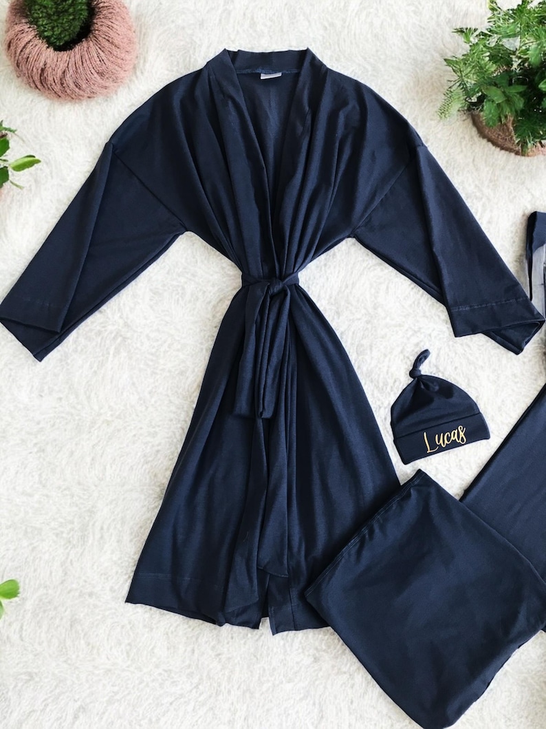 A flat-lay image featuring a navy blue maternity robe and pajama set with "Dad" written in gold script on a T-shirt, a matching robe, a baby hat with "Lucas" embroidered in gold, and a pair of pants, arranged on a soft white rug.