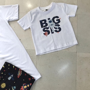 Family-themed clothing set featuring white T-shirts with space-themed "DAD," "BIG BRO," and "BIG SIS" text, a navy robe with colorful space designs, and a matching space-themed swaddle with a personalized hat. Laid out on a tiled floor.