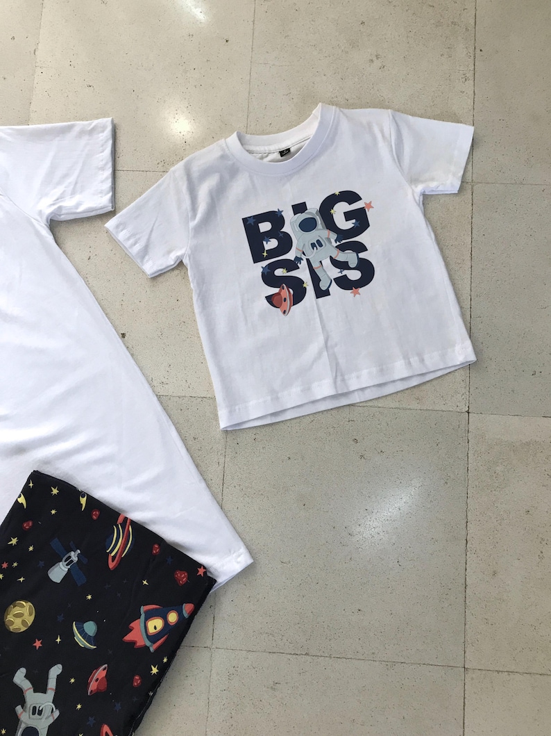 Family-themed clothing set featuring white T-shirts with space-themed "DAD," "BIG BRO," and "BIG SIS" text, a navy robe with colorful space designs, and a matching space-themed swaddle with a personalized hat. Laid out on a tiled floor.