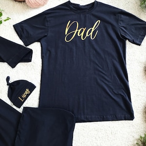A flat-lay image featuring a navy blue maternity robe and pajama set with "Dad" written in gold script on a T-shirt, a matching robe, a baby hat with "Lucas" embroidered in gold, and a pair of pants, arranged on a soft white rug.