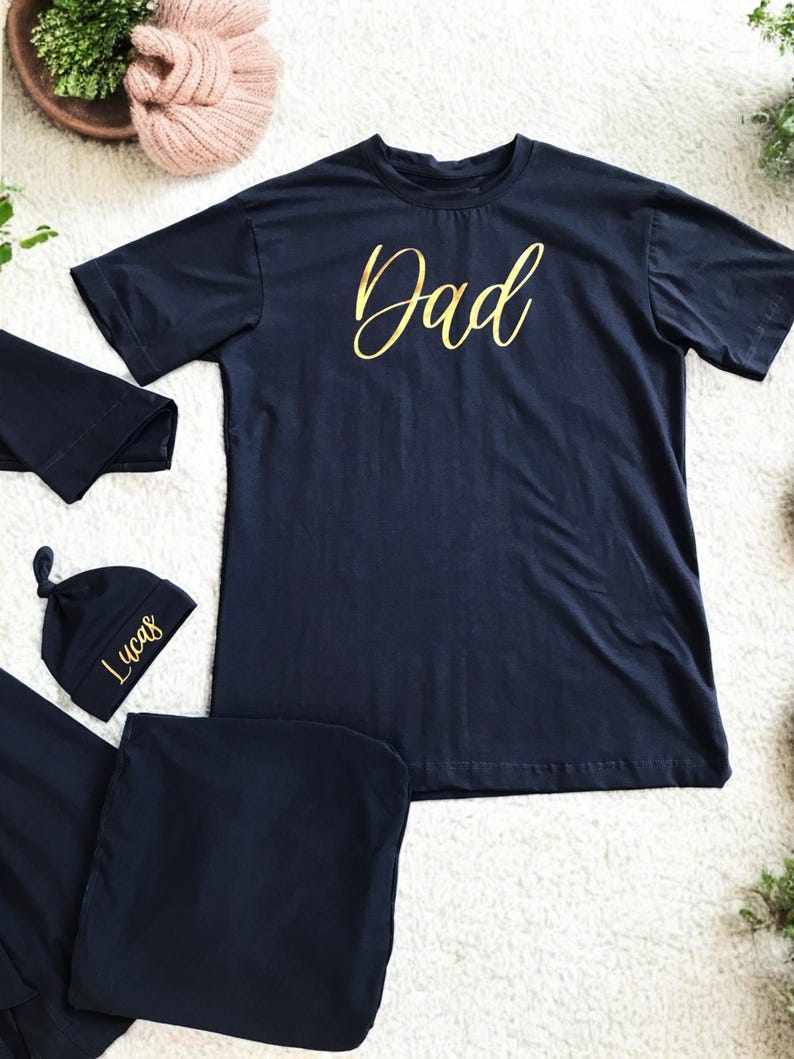 A flat-lay image featuring a navy blue maternity robe and pajama set with "Dad" written in gold script on a T-shirt, a matching robe, a baby hat with "Lucas" embroidered in gold, and a pair of pants, arranged on a soft white rug.