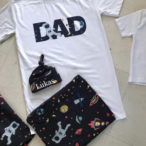 Family-themed clothing set featuring white T-shirts with space-themed "DAD," "BIG BRO," and "BIG SIS" text, a navy robe with colorful space designs, and a matching space-themed swaddle with a personalized hat. Laid out on a tiled floor.