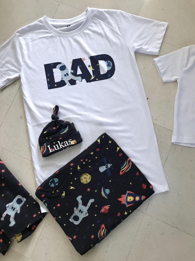 Family-themed clothing set featuring white T-shirts with space-themed "DAD," "BIG BRO," and "BIG SIS" text, a navy robe with colorful space designs, and a matching space-themed swaddle with a personalized hat. Laid out on a tiled floor.