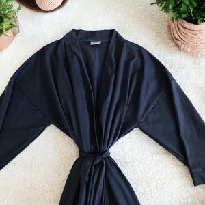 A flat-lay image featuring a navy blue maternity robe and pajama set with "Dad" written in gold script on a T-shirt, a matching robe, a baby hat with "Lucas" embroidered in gold, and a pair of pants, arranged on a soft white rug.