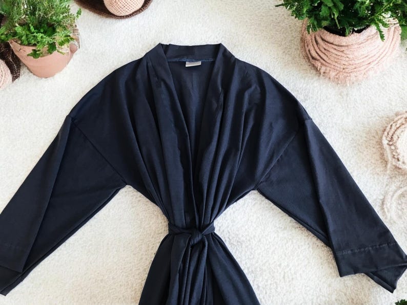 A flat-lay image featuring a navy blue maternity robe and pajama set with "Dad" written in gold script on a T-shirt, a matching robe, a baby hat with "Lucas" embroidered in gold, and a pair of pants, arranged on a soft white rug.
