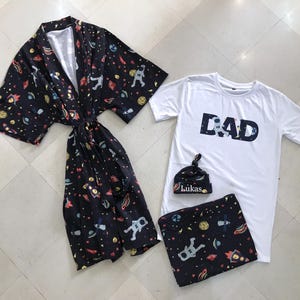 Family-themed clothing set featuring white T-shirts with space-themed "DAD," "BIG BRO," and "BIG SIS" text, a navy robe with colorful space designs, and a matching space-themed swaddle with a personalized hat. Laid out on a tiled floor.