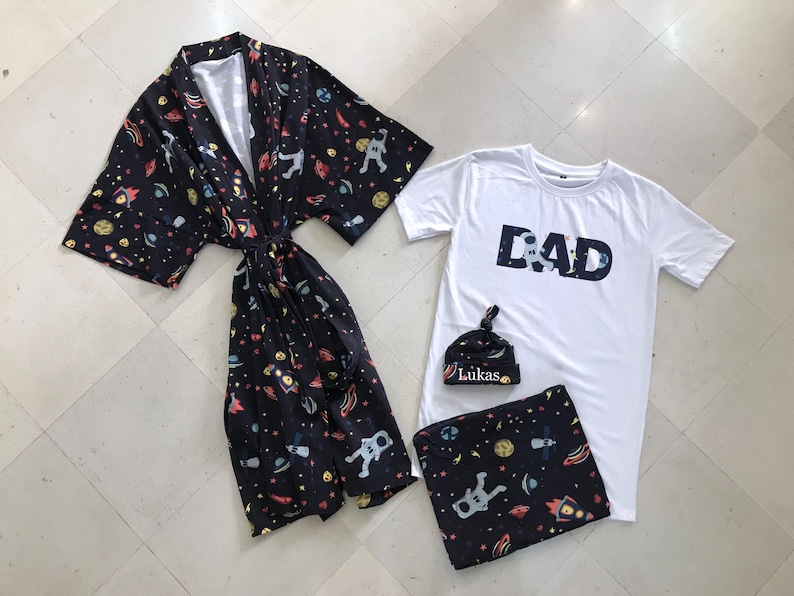 Family-themed clothing set featuring white T-shirts with space-themed "DAD," "BIG BRO," and "BIG SIS" text, a navy robe with colorful space designs, and a matching space-themed swaddle with a personalized hat. Laid out on a tiled floor.