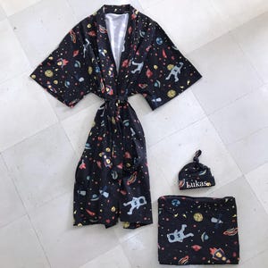 Family-themed clothing set featuring white T-shirts with space-themed "DAD," "BIG BRO," and "BIG SIS" text, a navy robe with colorful space designs, and a matching space-themed swaddle with a personalized hat. Laid out on a tiled floor.