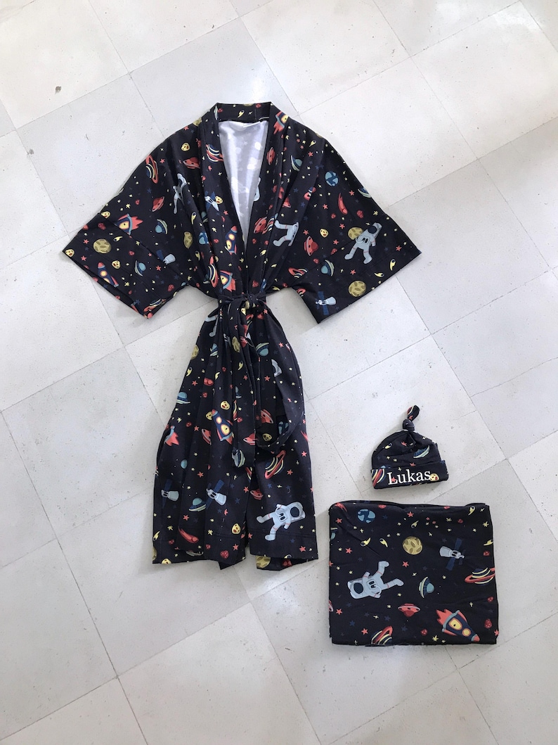 Family-themed clothing set featuring white T-shirts with space-themed "DAD," "BIG BRO," and "BIG SIS" text, a navy robe with colorful space designs, and a matching space-themed swaddle with a personalized hat. Laid out on a tiled floor.