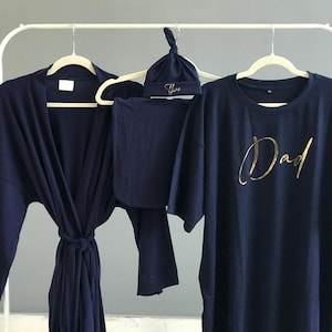 A flat-lay image featuring a navy blue maternity robe and pajama set with "Dad" written in gold script on a T-shirt, a matching robe, a baby hat with "Lucas" embroidered in gold, and a pair of pants, arranged on a soft white rug.