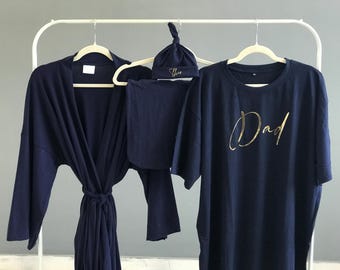 May include: Navy blue matching set of sleepwear and accessories. Includes a robe, a long-sleeve shirt with "Dad" in gold script, a hat, and a swaddle blanket. All items are hanging on a white rack.