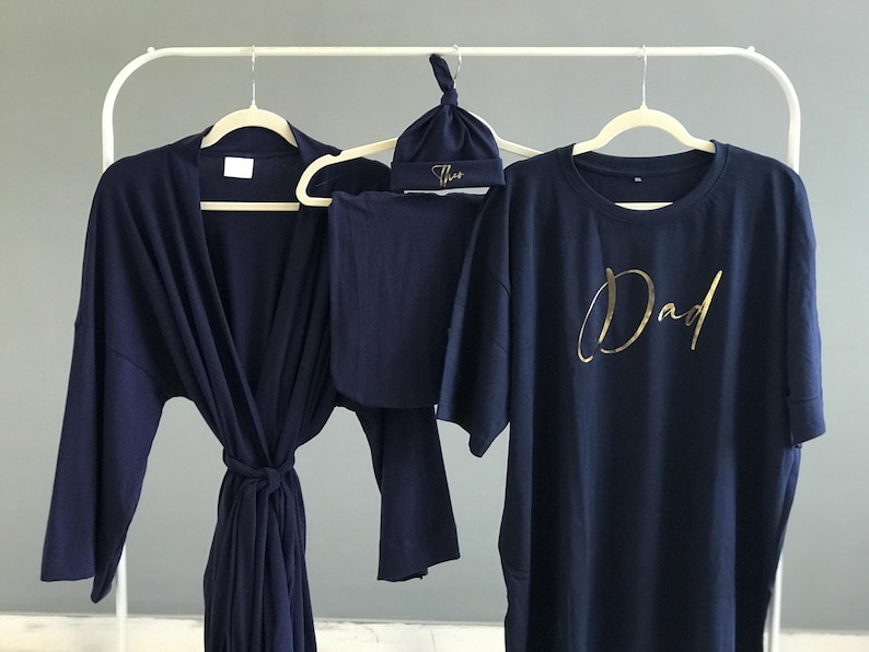 A flat-lay image featuring a navy blue maternity robe and pajama set with "Dad" written in gold script on a T-shirt, a matching robe, a baby hat with "Lucas" embroidered in gold, and a pair of pants, arranged on a soft white rug.