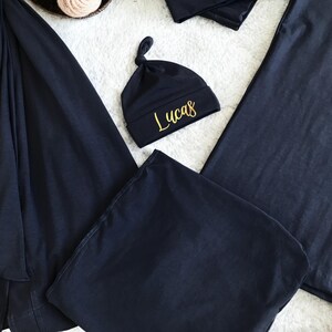 A flat-lay image featuring a navy blue maternity robe and pajama set with "Dad" written in gold script on a T-shirt, a matching robe, a baby hat with "Lucas" embroidered in gold, and a pair of pants, arranged on a soft white rug.