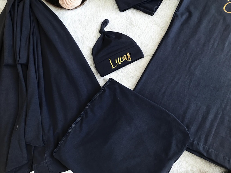 A flat-lay image featuring a navy blue maternity robe and pajama set with "Dad" written in gold script on a T-shirt, a matching robe, a baby hat with "Lucas" embroidered in gold, and a pair of pants, arranged on a soft white rug.