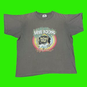 May include: A gray t-shirt with a cartoon character wearing a green hat and sunglasses. The character is holding a peace sign and a cigarette. The text "IRIE TOONS" and "Big up folks!" is printed on the shirt.