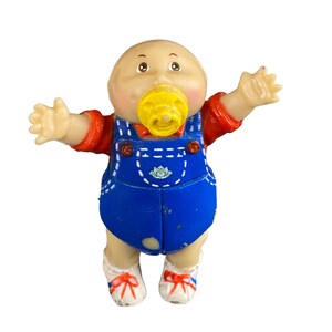 May include: A plastic Cabbage Patch Kid doll wearing blue overalls, a red shirt, and a yellow pacifier. The doll has a bald head and a rosy complexion.