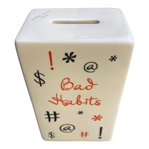 May include: White ceramic piggy bank with a slot on top. The bank has black and red symbols and the text "Bad Habits" on the front.