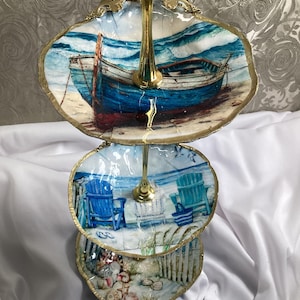 May include: A three-tiered serving tray with a gold-colored metal stand. Each tier features a coastal scene with a boat, beach chairs, and seashells. The edges of the trays are trimmed with gold. The tray is ideal for serving desserts or displaying decorative items.