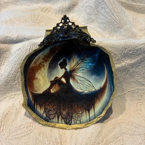 May include: A decorative shell-shaped trinket dish with a gold-painted edge and ornate metalwork. The interior features a fantasy scene of a fairy sitting on a crescent moon against a starry sky.