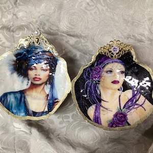 May include: Two decorative shell-shaped art pieces. One features a woman in a blue turban and dress, the other a woman in a purple head covering and dress. Both have gold trim and ornate embellishments.
