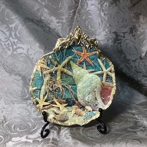 May include: Decorative seashell-shaped plaque with a gold-colored ornate frame. The plaque features a teal background with starfish, seashells, and a large conch shell. The edges are trimmed in gold, and the plaque is displayed on a black metal stand.