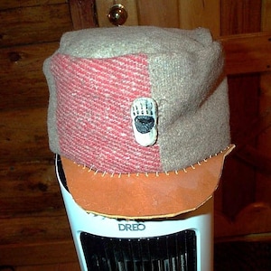 May include: A structured cap featuring a textured tan and red woven fabric crown with a prominent bear paw emblem centred on the front. The cap includes a smooth, light brown leather brim attached with visible white stitching along the base.