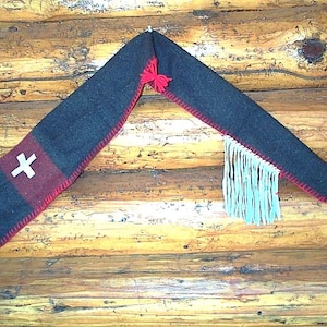 May include: A dark blue and red felt hat with a white cross design and red stitching. The hat has a long, pointed shape with a tassel at the end and a fringe detail. The hat is displayed against a wooden background.