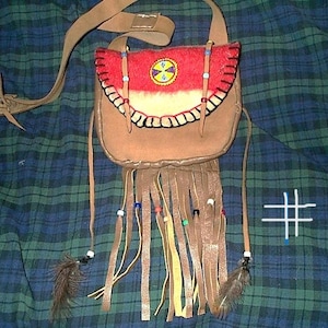 May include: A tan leather shoulder bag featuring a red flap with a circular yellow and blue beaded emblem. The bag includes long leather fringe strips adorned with small colorful beads and two brown feathers. The number #1 is written on the right.