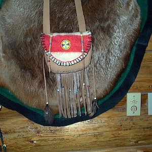 May include: A small, tan leather shoulder bag with a red and white patterned flap. The bag features fringe, beads, and a decorative emblem. The bag is suspended against a brown fur-like background.