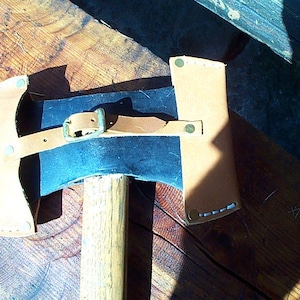 Puede incluir: Un hacha de doble filo con mango de madera y funda de cuero. La cabeza del hacha es azul oscuro y plateada, con una funda de cuero color canela asegurada por una correa y hebilla de cuero marrón. El hacha descansa sobre una superficie de madera.