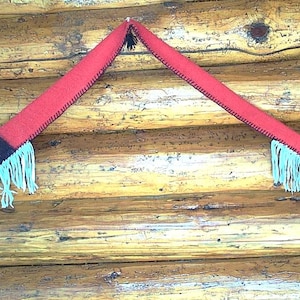 May include: A long, red felted scarf featuring a dark brown rectangular accent, light blue fringe detailing, and black whip-stitch edging. The scarf includes small black tassels at the ends and is draped in an inverted V-shape, showcasing its textured fabric and decorative construction.