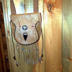 May include: A tan leather pouch with a long shoulder strap, featuring dark brown whipstitching along the edges, a central black stone inlay, a circular beaded medallion, and long leather fringe adorned with multi-colored beads and feathers.