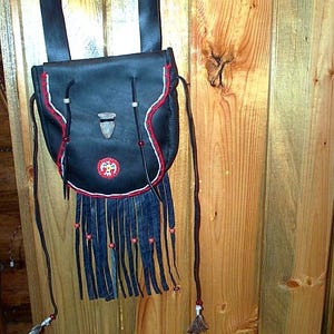 May include: A black leather pouch with a long shoulder strap, featuring a flap closure secured by a stone-like toggle. The bag is decorated with red and white stitching, a circular beaded emblem, and long leather fringe with small beads.