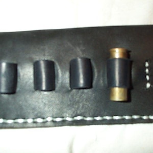May include: A black leather cartridge holder featuring four individual loops for ammunition storage. The rectangular piece is constructed with visible white stitching along the bottom edge and sides, with one brass-colored metal cartridge inserted.