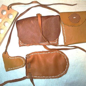 May include: A collection of handmade leather pouches and a wooden block with holes. The pouches vary in size and shape, with visible white stitching and leather straps. The wooden block is rectangular with multiple circular openings.