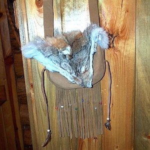 May include: A tan suede shoulder bag with a coyote fur accent. The bag features a fringe bottom with decorative beads and feathers. The bag hangs against a wooden wall.